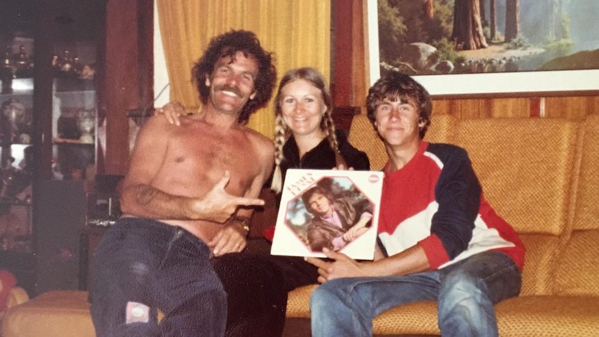 A shirtless moustached man points at an LP record on an old couch next to a woman with piggy tails and young man.