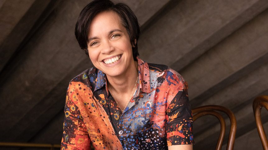 Jada Alberts smiling broadly wearing a colourful Indigenous-print shirt, short brown hair